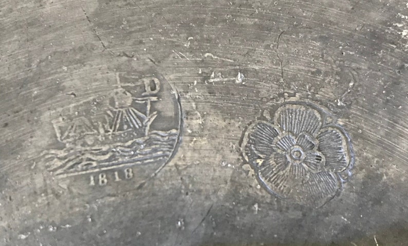 May include: Close-up of a pewter plate with two embossed designs. The left design features a ship with the date 1818 below it. The right design is a stylized flower.