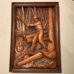 Può includere: Una scultura in legno a rilievo che raffigura un uomo che abbatte un albero in un ambiente boschivo. La scultura è incorniciata in una cornice di legno scuro.
