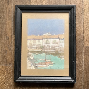 May include: A framed watercolor painting depicting a harbor scene with several boats docked in front of a row of white buildings. The sky is a light blue with hints of purple and pink. The water is a vibrant turquoise.