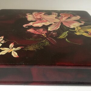 May include: A dark red wooden box with a hinged lid. The lid is decorated with a hand-painted floral design featuring pink, white, and yellow flowers.
