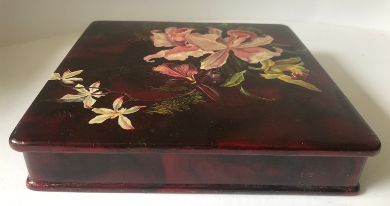 May include: A dark red wooden box with a hinged lid. The lid is decorated with a hand-painted floral design featuring pink, white, and yellow flowers.