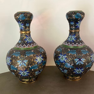 Peut inclure: Deux vases décoratifs avec un corps bulbeux et un col fin. Les vases présentent un fond noir avec un motif floral bleu, or et vert. Les bords et les bases sont garnis d'or.