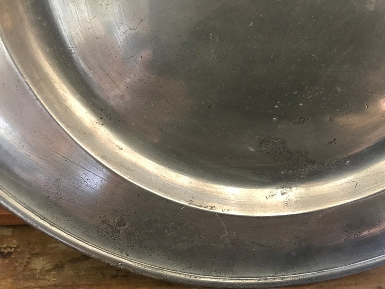 May include: A silver-colored metal plate with a slightly raised rim. The plate has a smooth, shiny surface with some scratches and marks.