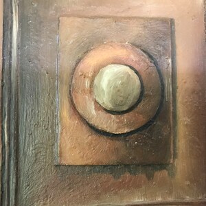 Op de afbeelding: Een close-up van een houten deur met een geschilderd detail van een cirkel in een vierkant. De cirkel is bruin met een kleinere, cr&egrave;mekleurige cirkel in het midden.