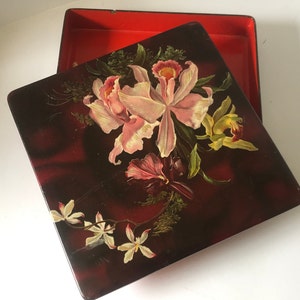 May include: A square wooden box with a hinged lid. The lid is decorated with a floral design featuring pink and yellow orchids on a dark red background.
