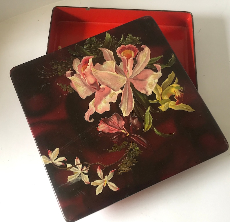 May include: A square wooden box with a hinged lid. The lid is decorated with a floral design featuring pink and yellow orchids on a dark red background.