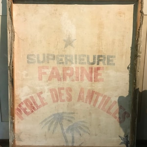 May include: A vintage framed canvas sign with the text "SUPERIEURE FARINE PERLE DES ANTILLES" in red and blue lettering. The sign features a star and two palm trees in blue and white.