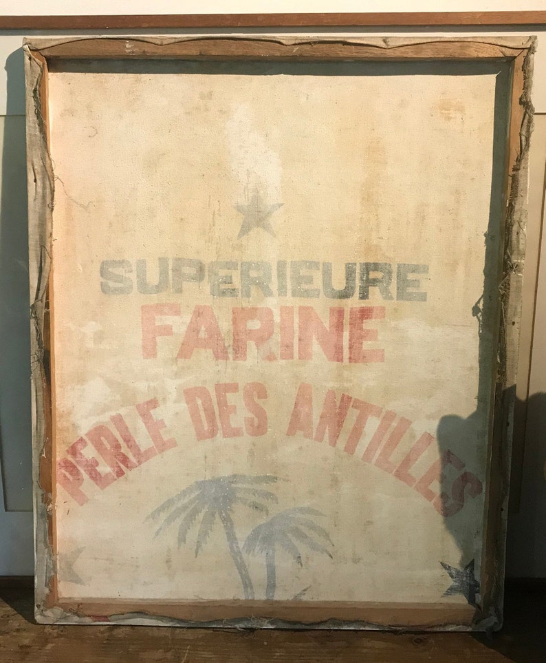 May include: A vintage framed canvas sign with the text "SUPERIEURE FARINE PERLE DES ANTILLES" in red and blue lettering. The sign features a star and two palm trees in blue and white.