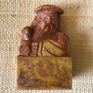 Puede incluir: Una figurita de madera tallada de una deidad china, con una larga barba y un sombrero, sosteniendo un bastón. La figurita está sentada sobre una base cuadrada con un diseño tallado de un paisaje.