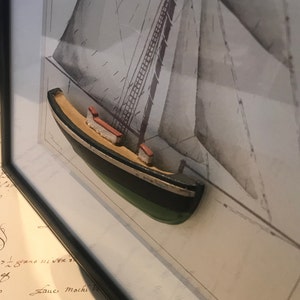 May include: A framed print of a sailboat with a 3D model of a small boat in the foreground. The model boat is painted black, green, and white.