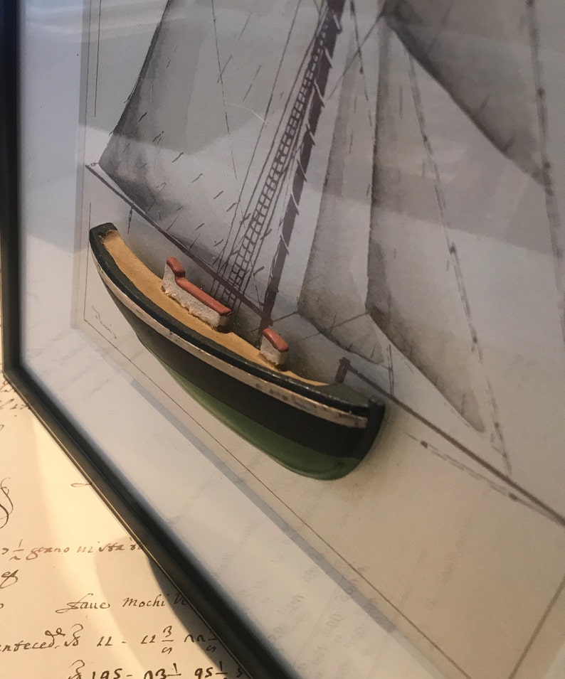 May include: A framed print of a sailboat with a 3D model of a small boat in the foreground. The model boat is painted black, green, and white.