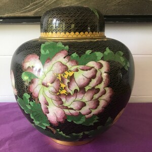 Op de afbeelding: Een zwarte cloisonné pot met een deksel, versierd met een bloemenmotief van roze, groene en gele bloemen. De pot heeft een gouden rand en een zwarte achtergrond.