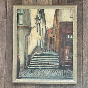 Może przedstawiać: Oprawiony obraz olejny przedstawiający europejską scenę uliczną. Brukowana ulica prowadzi w górę po schodach, po bokach budynki i latarnia uliczna. Osoba stoi po lewej stronie obrazu, widoczny jest napis "LA".