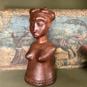 May include: A dark brown wooden sculpture of a woman's bust with a detailed carved face and a simple necklace. The sculpture is set against a painted background.