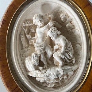 May include: An oval-shaped wall plaque featuring a cream-colored relief carving of two children and a lamb in a landscape setting. The central figures are surrounded by a thin gold-toned metal frame set within a polished wooden border.