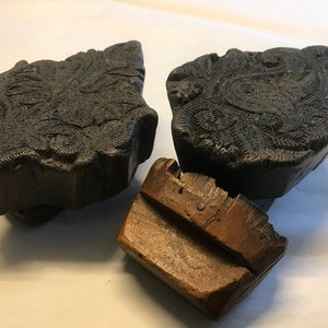 May include: Three vintage wooden printing blocks with intricate carved designs. The blocks are dark brown and black and have a worn, antique look.