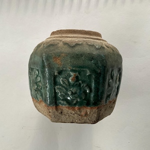 Può includere: Un piccolo vaso antico in ceramica verde con superficie strutturata e motivi floreali in rilievo. Il vaso ha un corpo arrotondato, un collo corto e un aspetto invecchiato. La base è di colore marrone-rossastro.