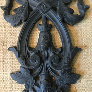 May include: A black metal decorative wall hanging with intricate floral designs. The design features a central oval shape with a smaller oval inside. The piece has a distressed finish.