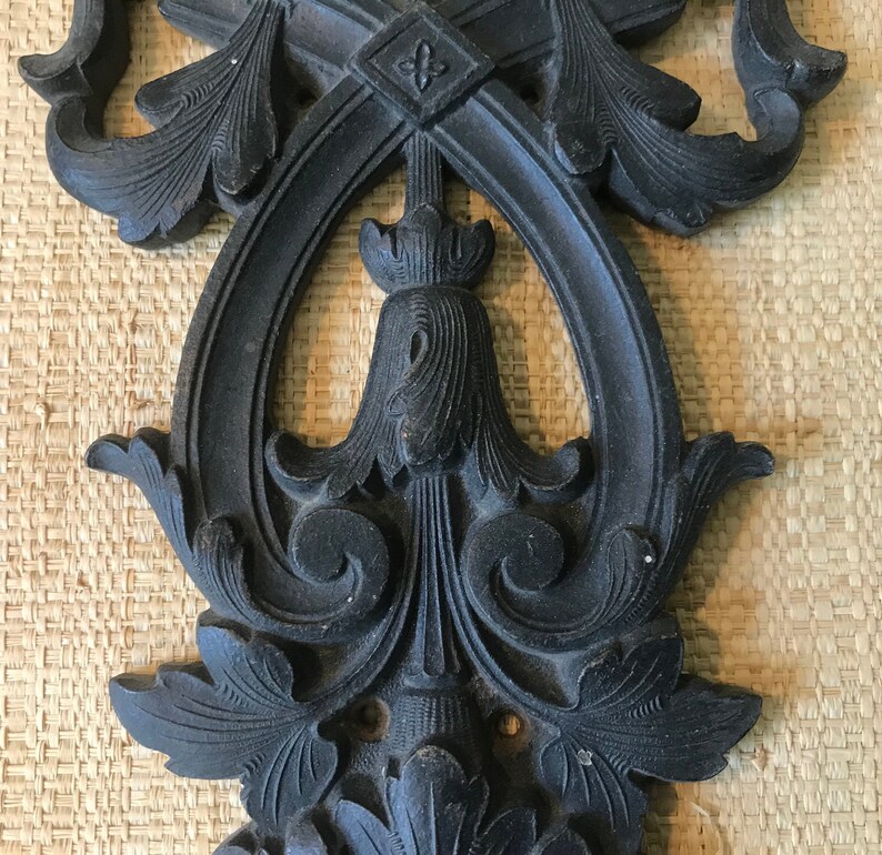 May include: A black metal decorative wall hanging with intricate floral designs. The design features a central oval shape with a smaller oval inside. The piece has a distressed finish.