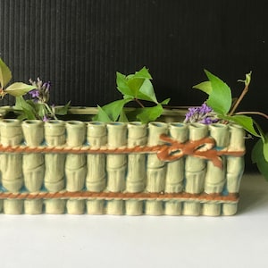 May include: A green ceramic planter shaped like a bamboo fence with brown rope accents. The planter has multiple holes for planting. The planter is filled with green vines and purple flowers.