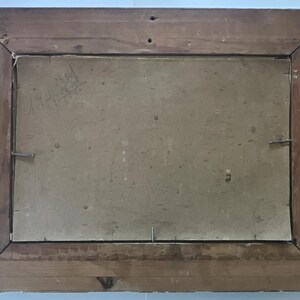 May include: Back of a framed painting. The wooden frame is a medium brown color with visible wood grain. The backing is a light tan color, secured with metal staples and a single nail.
