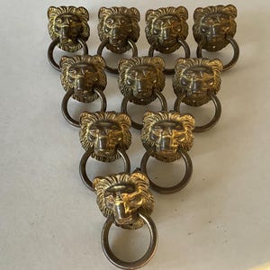 Peut inclure: Dix poignées de tiroir en laiton en forme de tête de lion avec des anneaux. Les têtes de lion ont des crinières et des traits du visage détaillés. Les poignées sont disposées en triangle.