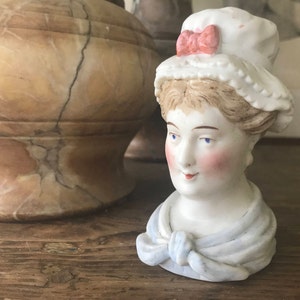 Może przedstawiać: Porcelanowa figurka głowy kobiety w białym czepeczku z różową kokardką i niebieskim szalem. Figurka ma namalowane niebieskie oczy, różowe policzki i uśmiech.