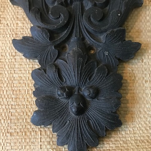 May include: A black metal decorative wall plaque with a floral design and a face in the center.