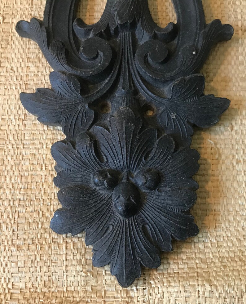 May include: A black metal decorative wall plaque with a floral design and a face in the center.