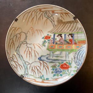 Op de afbeelding: Een ronde, antieke porseleinen schaal met een handgeschilderde scène. De schaal toont een gedetailleerde illustratie van twee figuren in een pagode, omgeven door een landschap van bomen, bloemen en een rivier. Het kleurenpalet omvat rood, blauw, groen en bruin.