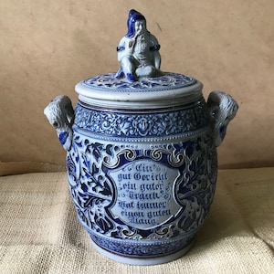 Peut inclure: Un pot en céramique bleu et blanc avec un couvercle et deux poignées. Le pot est décoré de motifs floraux et d'une petite figurine d'un homme avec une barbe et un chapeau. Le pot porte l'inscription "Ein gut Gerich't ein guter Trunk hat immer einen guten Klang" sur le devant.