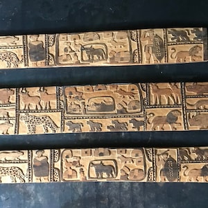 Può includere: Tre pannelli in legno con immagini intagliate di vari animali africani, tra cui una girafa, un leone, un leopardo e un rinoceronte. I pannelli sono disposti orizzontalmente, con il pannello superiore che mostra una figura umana che tiene uno scudo.