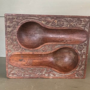 Puede incluir: Un reposacucharas rectangular de madera con dos huecos en forma de cuchara. El borde está decorado con motivos florales y de hojas talladas. La madera tiene un tono marrón cálido con vetas visibles y signos de desgaste.