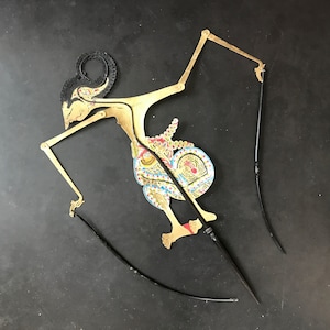 Puede incluir: Un títere de sombras tradicional indonesio, o wayang kulit, que representa una figura con un traje colorido y estampado. El títere está unido a un conjunto de varillas negras para su manipulación.