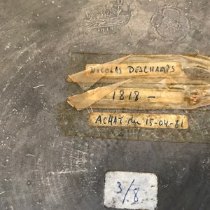 May include: A close-up of a gray metal surface with handwritten text on three labels. The labels read "Nicolas Deschamps", "1818", and "Achat du 15-04-81". There is also a smaller label with the number "3/8" written on it.
