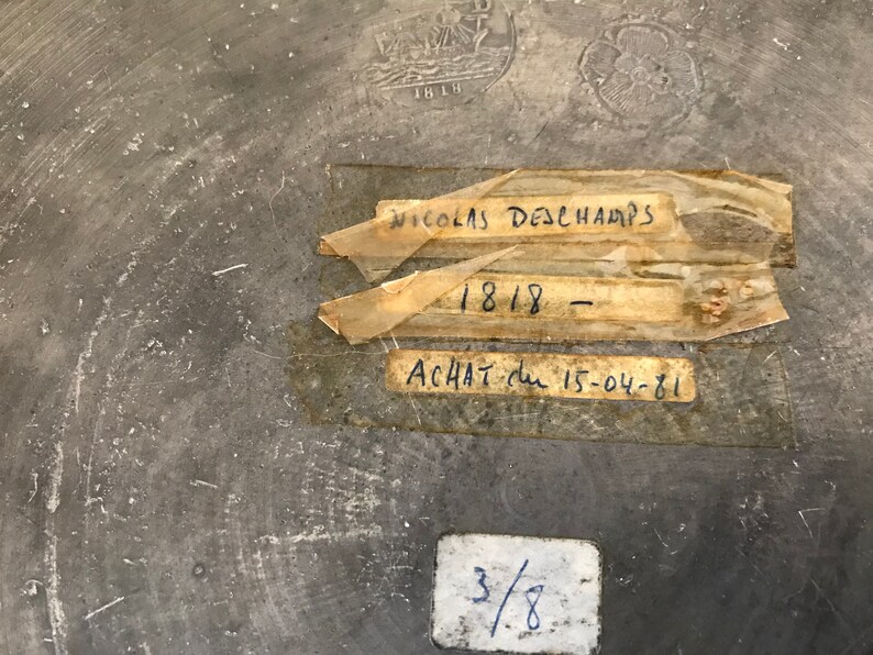 May include: A close-up of a gray metal surface with handwritten text on three labels. The labels read "Nicolas Deschamps", "1818", and "Achat du 15-04-81". There is also a smaller label with the number "3/8" written on it.
