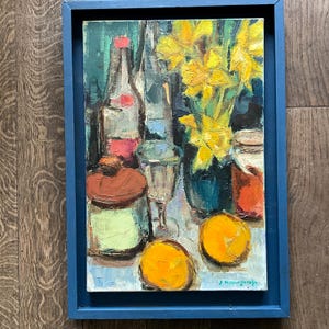 May include: Oil painting in a blue frame depicts a still life with a wine bottle, glass, jar, and yellow flowers. Two oranges sit in the foreground. The painting has a textured, impressionistic style.