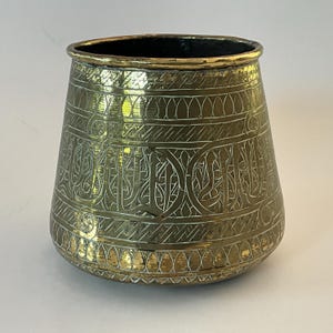May include: A brass planter with intricate engraved designs, including Arabic calligraphy. The planter has a wide, rounded base and a flared top.