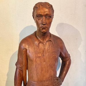 Può includere: Una scultura in legno di una persona che tiene un bastone. La scultura è scolpita in uno stile realistico e ha un colore marrone caldo.