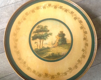 May include: A round, wooden tray with a green border and a gold painted design. The center of the tray features a painted scene of a castle in a landscape with trees.
