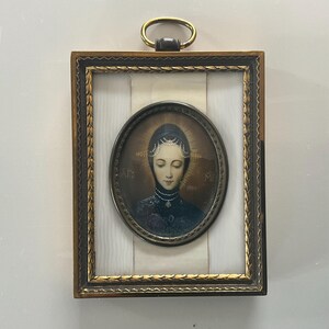 Op de afbeelding: Een antiek religieus icoon met een portret van een vrouw met een halo, omlijst in een goudkleurige rechthoekige lijst. Het ovale portret staat tegen een witte achtergrond, met een gouden ring aan de bovenkant om op te hangen.