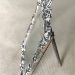 May include: Silver-toned ornate picture frame with a glass insert and a stand. The frame has a detailed design with floral motifs.