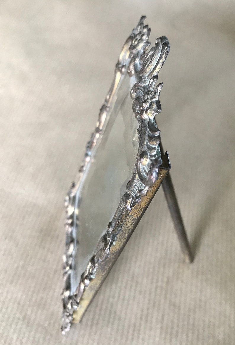 May include: Silver-toned ornate picture frame with a glass insert and a stand. The frame has a detailed design with floral motifs.