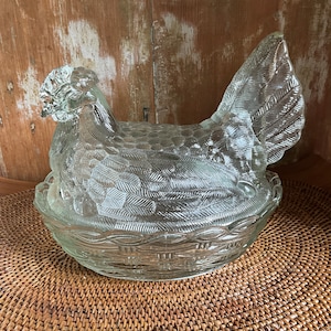 May include: A clear glass hen-shaped dish with a textured surface. The hen sits atop a basket-weave style base. The dish is designed to hold eggs or other items. The glass has a detailed feather pattern.