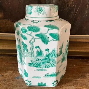 Peut inclure: Un pot en porcelaine blanche avec un motif floral vert et blanc sur le couvercle et une scène verte et blanche de personnes dans un jardin sur le corps. Le pot a une forme hexagonale.