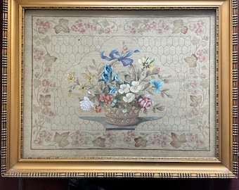 May include: A framed floral tapestry featuring a basket overflowing with colorful flowers, including blue, white, and pink blooms. The tapestry is set against a beige background with a lattice pattern and framed in a gold-colored wooden frame.