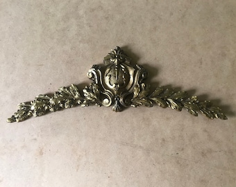 May include: A gold-toned metal decorative trim with a central ornate design and leaf-like accents on either side.