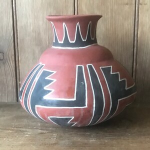 Könnte beinhalten: Eine rote Tonvase mit schwarzen und weißen geometrischen Mustern. Die Vase hat einen runden Körper und einen schmalen Hals.