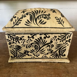 May include: A rectangular box with a matching domed lid, featuring a cream-coloured base with a hand-painted black floral and leafy pattern. The design covers the front and top surfaces, showcasing intricate, flowing botanical motifs in black.