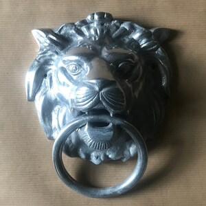 Op de afbeelding: Een zilveren deurklopper in de vorm van een leeuwenkop met een ronde ring. Het gezicht van de leeuw is gedetailleerd met een manen, neus en ogen.